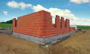 Budowa domu 100m2 w toku. Czerwone ściany z pustaków, fundamenty z bloczków betonowych, niebo z chmurami.