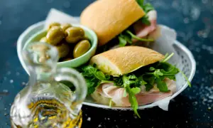 Två halvor av en ciabatta macka med prosciutto och ruccola, serverade med gröna oliver och en karaff olivolja.