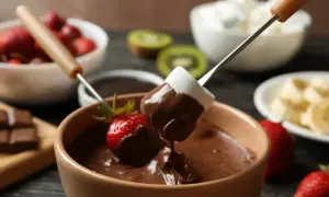 Czekoladowe fondue z pianką i truskawką na patyczku, w tle owoce i pianki.