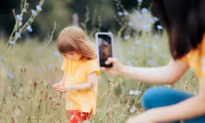 Dziecko w żółtej koszulce zbiera kwiatki na łące, a mama dokumentuje chwilę telefonem, praktykując sharenting.