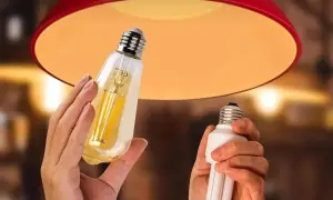 Une ampoule connectée orange, au design vintage, est tenue à côté d'une ampoule fluocompacte, sous un abat-jour rouge.
