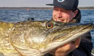 Glücklicher Angler hält stolz einen großen Hecht, gefangen nach einem Gewitter. Das Wasser ist ruhig, die Sonne scheint.