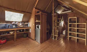 Intérieur d'une cabane en bois avec coin cuisine, réfrigérateur, étagères et échelle menant à un lit. Un havre de paix en Haute-Loire.