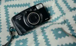 Czarny aparat Yashica Microtec Zoom 90 z paskiem, leżący na białym materiale z turkusowymi zygzakami. Wygląda jak z lat 90.