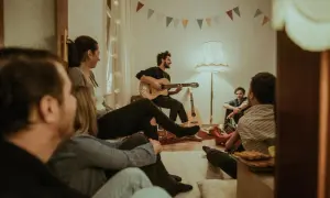 Freunde versammeln sich für einen gemütlichen Abend mit Gitarrenmusik und **Lieder zum Mitsingen**. Bunte Wimpelketten schmücken die Wand.
