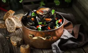 Un grand pot en cuivre rempli de moules ouvertes avant cuisson, garnies de persil et de piment. Du pain rustique est disposé sur la table en bois.