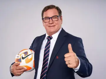 Uśmiechnięty trener reprezentacji Polski w garniturze trzyma piłkę i pokazuje kciuk w górę.