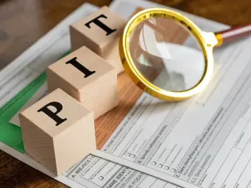 Drewniane klocki z literami P, I, T układają się w słowo "PIT". Obok leży lupa i formularz podatkowy.