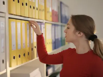 Kobieta w czerwonym swetrze sięga po żółty segregator, porządkując dokumenty. To część jej zakresu obowiązków.