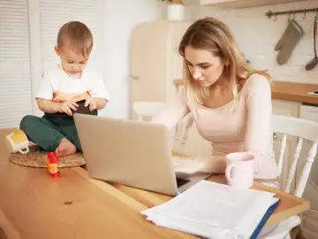 Młoda mama pracuje na laptopie, podczas gdy jej małe dziecko bawi się obok. Czy na urlopie macierzyńskim można pracować? Tak, często jest to możliwe.