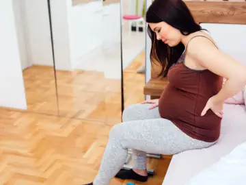 Kobieta w 27 tygodniu ciąży siedzi na łóżku, masując bolący odcinek lędźwiowy.