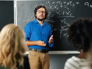 Nauczyciel w niebieskiej koszuli tłumaczy matematykę przy tablicy. Czy nauczyciel jest funkcjonariuszem publicznym? To pytanie nurtuje wielu.