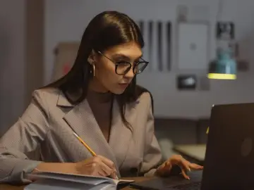 Kobieta w okularach pracuje przy laptopie, notując w zeszycie. Zastanawia się nad limitem nadgodzin w roku, by zachować równowagę.
