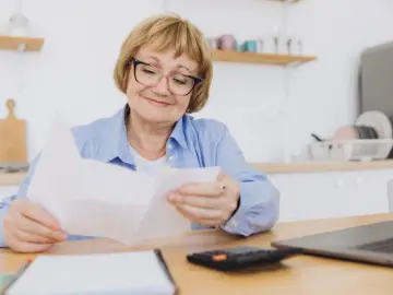 Starsza pani z okularami przegląda dokumenty. Może to rachunki z rp-7 zlikwidowanego zakładu pracy.