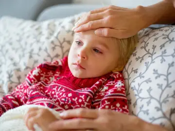 Małe dziecko w czerwonym sweterku z wzorem płatków śniegu leży chore, z gorączką. Ręka opiekuna sprawdza temperaturę. To typowy obrazek, gdy dopada nas wirus.