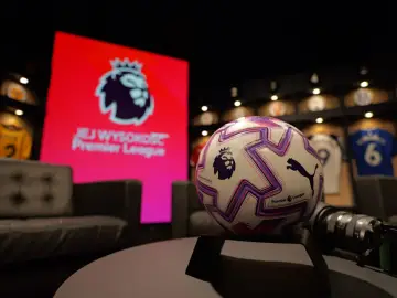 Piłka do gry w Premier League na tle ekranu z logo ligi. W tle widoczne koszulki piłkarskie.