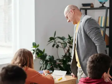 Nauczyciel pochyla się nad uczennicą, która pisze. W tle roślina i regał z książkami. Czy to początek choroby zawodowej nauczycieli?