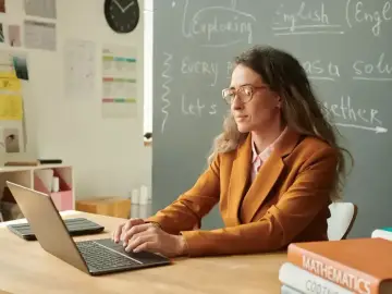 Nauczycielka pracuje przy laptopie. Na tablicy widnieje napis "English". Karta oceny pracy nauczyciela może być wypełniana cyfrowo.