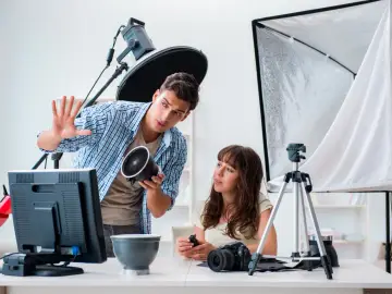 Mężczyzna, technik fotografii i multimediów, pokazuje kobiecie lampę. W tle sprzęt studyjny.