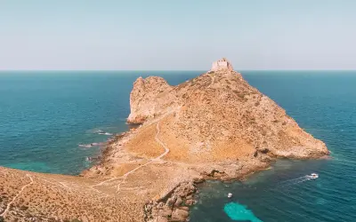 Rocciosa isola con un antico fortino in cima, circondata da acque turchesi. Un sentiero serpeggia verso la vetta, evocando le spiagge delle isole Egadi.