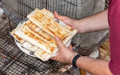 Talerz z grillowanymi wrapami kebab, trzymany przez ręce na tle rusztu i kamieni.