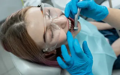 Pacjentka w okularach ochronnych przechodzi leczenie kanałowe. Dentysta w niebieskich rękawiczkach używa wiertła, by ulżyć w bólu zęba.