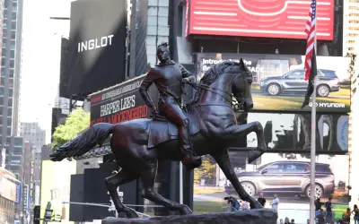 A bronze statue of a person on horseback stands in Times Square, with billboards for Inglot and Harper Lee's "To Kill a Mockingbird" in the background.