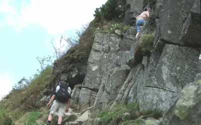 Turysta z plecakiem wspina się po skalistej ścieżce, obok wspina się inna osoba.