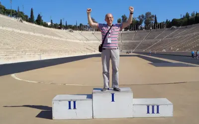 Mężczyzna na podium w historycznym stadionie panatenajskim, z rękami w górze, świętuje zwycięstwo.