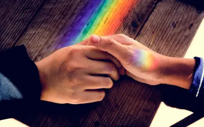 Dłonie splecione na drewnianym blacie, na których widać tęczowe refleksy. To jak pierścionek nastroju znaczenie kolorów, symbolizujący różnorodność i nadzieję.