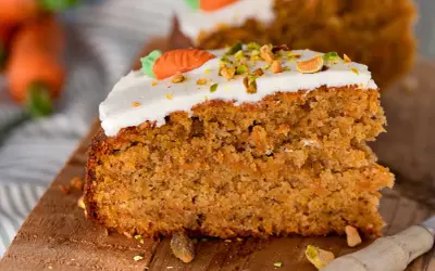 Ein saftiges Stück Karottenkuchen aus der Kastenform, dekoriert mit weißem Zuckerguss, gehackten Pistazien und einer kleinen Marzipankarotte.