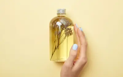 Eine Hand mit hellblauen Nägeln berührt eine Flasche mit gelbem Öl und getrockneten Blumen. Perfekt für alle, die Duschgel selber machen wollen.