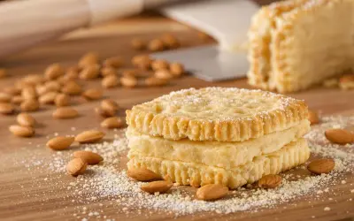Trois biscuits joconde empilés, saupoudrés de sucre glace, entourés d'amandes sur une planche en bois.