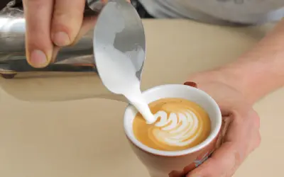 Barista gießt cremigen Milchschaum in eine Tasse Kaffee. Der Cappuccino Milchschaum bildet ein schönes Muster.