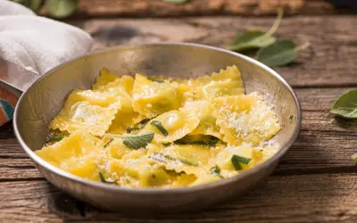 Ravioli ricotta e spinaci dorati, conditi con parmigiano e foglie di salvia fresca, serviti in una ciotola metallica su un tavolo di legno rustico.