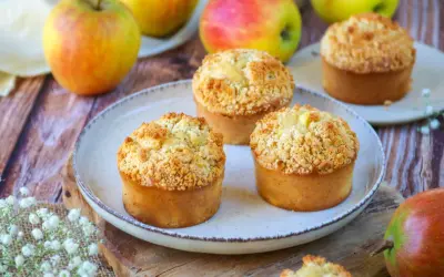 Trois muffins pomme amande dorés avec un crumble croustillant, entourés de pommes fraîches sur une table en bois.