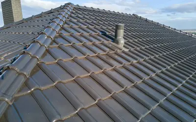Szary dach z dachówki ceramicznej, z widocznym kominkiem wentylacyjnym i kominem. Rozmieszczenie kominków wentylacyjnych na dachu zapewnia prawidłową cyrkulację powietrza.