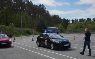 Na placu manewrowym, gdzie obowiązuje ruch lewostronny, czarny samochód szkoleniowy i czerwony pojazd czekają na kursantów. W tle niebieski autokar.