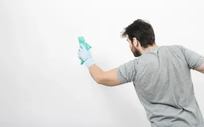 Un homme en gant bleu nettoie un mur blanc avec un chiffon vert. Il montre comment nettoyer un mur blanc sans enlever la peinture.