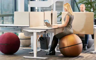 Kvinna arbetar vid ett höj- och sänkbart skrivbord i ett aktivitetsbaserat kontor. Hon sitter på en stor, brun träningsboll.