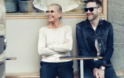 Joakim Berg familj: en kvinna och en man sitter på en bänk utanför ett café, båda ler och bär solglasögon.