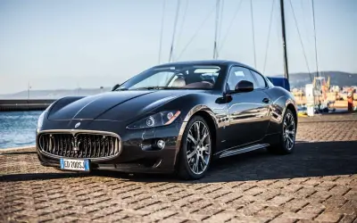 Eleganckie, czarne coupe auto Maserati stoi na nabrzeżu, z żaglami łodzi w tle.