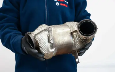 Osoba w rękawiczkach trzyma metalowy element, prawdopodobnie **fap** (filtr cząstek stałych) do samochodu.