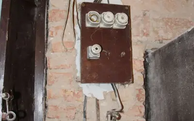 Stara instalacja elektryczna z ceramicznymi bezpiecznikami i widocznymi przewodami na ceglanej ścianie.