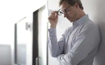 Mann mit Brille, der sich an eine Wand lehnt. Er wirkt nachdenklich, vielleicht kämpft er mit Hitzewallungen.