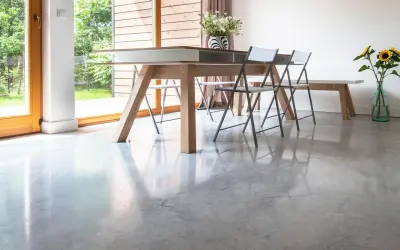 Jasny, nowoczesny salon z dużymi oknami i podłogą z polerowanego betonu. Stół z krzesłami i wazonem kwiatów.