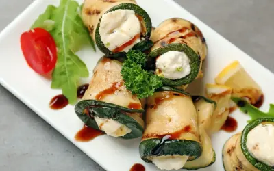 Deliziosi involtini di zucchine grigliate ripieni di formaggio cremoso, guarniti con pomodorini, rucola e salsa scura.