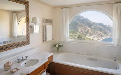 Un bagno di lusso con vasca, lavandino e specchio decorato, che offre una vista mozzafiato sulle montagne e sul mare. Tra i bagni più belli ed eleganti.
