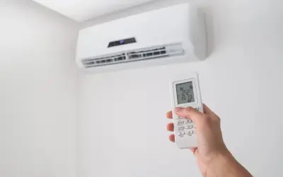 Dłoń trzymająca pilot do klimatyzacji na tle białej ściany z jednostką klimatyzacyjną.
