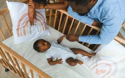Parents hold their baby's hand in a crib with a breathable mattress, a safe sleep environment.